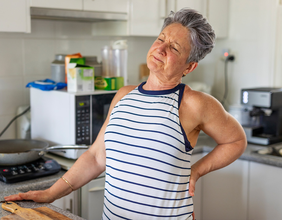 an elderly woman stretching in a kitchen with back pain