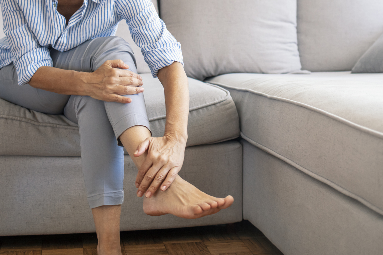 A woman in a blue & white striped shirt & gray pants sits on her gray couch clutching her ankle