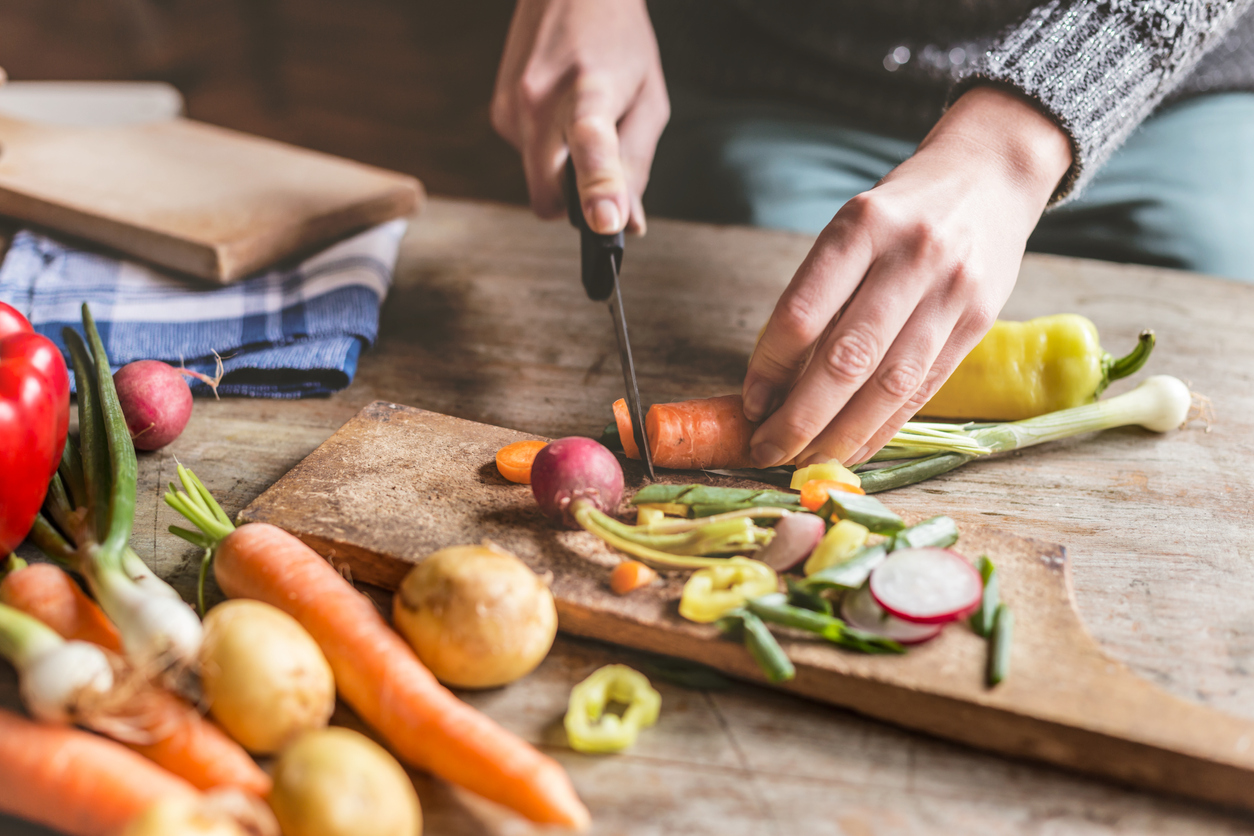 Closeup of a woman’s hands chopping a carrot while potatoes & peppers sit nearby